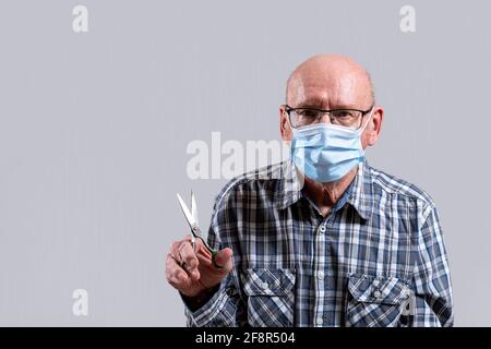 Vecchio calvo con occhiali e maschera medica con forbici in acciaio in mano. Spazio di copia. Vista frontale, scatto orizzontale interno. Foto Stock