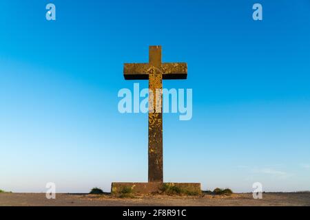 Grande vecchia croce di pietra cristiana coperta di lichene giallo luminoso di fronte ad un cielo blu chiaro all'inizio del tramonto. Concetto di fede e religione. Foto Stock