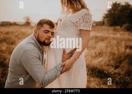 marito abbracci moglie incinta padre ascolta il ventre messo il suo orecchio al tummy. Felice famiglia riposarsi in natura abbracci in estate al tramonto. Futura madre CA Foto Stock