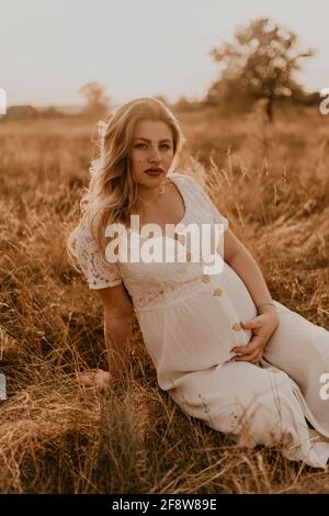 Giovane donna bionda caucasica incinta in abito di lino bianco di cotone siede e si trova nel mezzo del prato su erba secca in estate al tramonto in natura Foto Stock