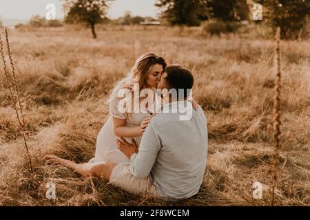 la moglie incinta si siede tra le braccia del marito e lo bacia e lo abbraccia. Felice famiglia riposo natura abbracci baci in estate al tramonto. Futura madre caucasica w Foto Stock