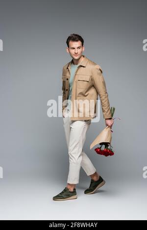 lunghezza completa di uomo trendy che tiene bouquet di rose rosse avvolto in carta mentre si cammina su grigio Foto Stock