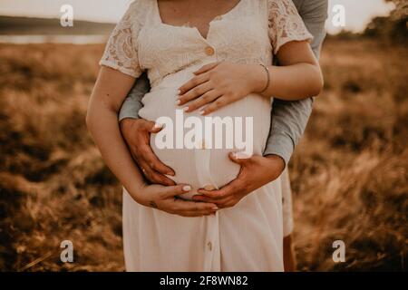 mani di futura madre e padre aggrappato tummy grande incinta. marito abbraccia moglie incinta. Famiglia felice riposarsi in natura abbracci in estate al tramonto. CAU Foto Stock