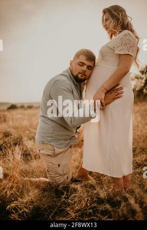 marito abbracci moglie incinta padre ascolta il ventre messo il suo orecchio al tummy. Felice famiglia riposarsi in natura abbracci in estate al tramonto. Futura madre CA Foto Stock