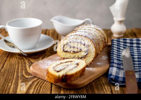 Rotolo di biscotto non tagliato con marmellata di frutta e zucchero in polvere su un tagliere, con una tazza di tè bianco. Cottura fatta in casa. Colazione. Foto Stock