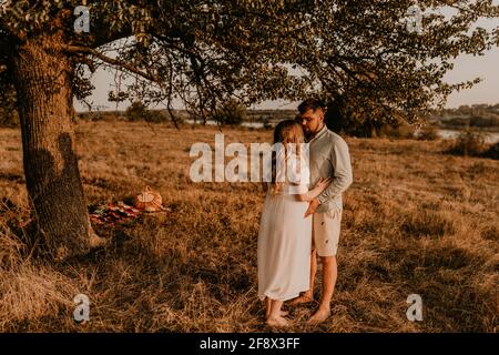 marito abbraccia la moglie incinta con il pancia grande. Felice famiglia riposante in natura abbracci in estate al tramonto. Coppia sullo sfondo di grande albero solitario. Foto Stock