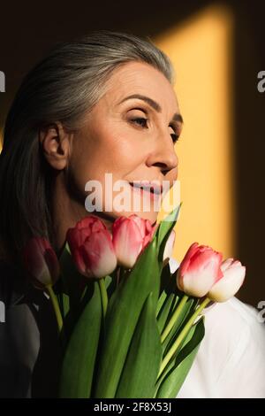 Donna di mezza età che tiene tulipani rosa su sfondo giallo con ombra Foto Stock