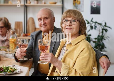 Ritratto di coppia anziana che guarda la fotocamera mentre si siede a. tavolo da pranzo e vino da bere durante la cena Foto Stock