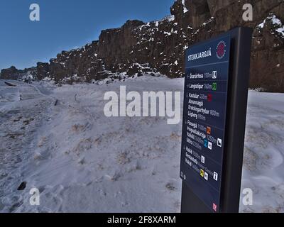 Vista del cartello con destinazioni e distanze accanto al sentiero innevato nel Parco Nazionale di Thingvellir nella stagione invernale con rocce vulcaniche. Foto Stock