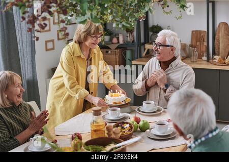 Donna anziana che serve torta di compleanno per l'uomo anziano mentre loro seduto al tavolo con gli amici a casa Foto Stock