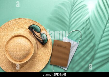Cappello di paglia, occhiali da sole, passaporto e maschera medica su sfondo verde con le foglie di palma ombra. Vacanze estive durante l'epidemia di coronavirus. Foto Stock