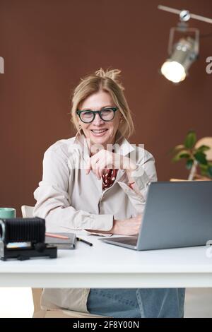 Ritratto di uomo d'affari maturo in occhiali sorridendo a macchina fotografica mentre seduto sul posto di lavoro con un computer portatile Foto Stock