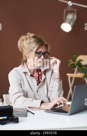 Donna d'affari matura in occhiali seduto al tavolo e digitando su un computer portatile in ufficio Foto Stock