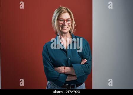 Ritratto di felice donna matura in occhiali sorridenti alla macchina fotografica mentre si è in piedi con le braccia incrociate contro lo sfondo rosso Foto Stock