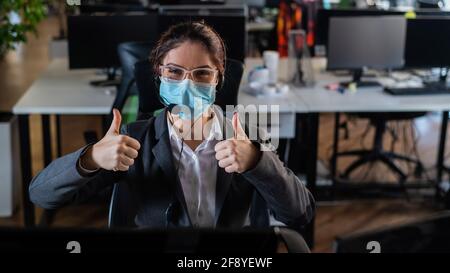 Donna caucasica in maschera medica mostrando pollici in ufficio. Foto Stock