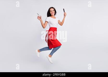 Foto profilo full size di bella bruna ottimista capelli lunghi donna con forbici salto indossare t-shirt bianca isolato su grigio sfondo a colori Foto Stock