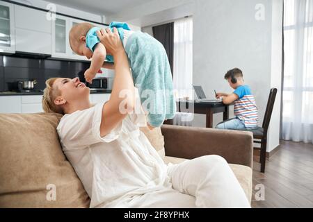 Felice madre che gioca con il suo figlio più giovane sul divano mentre la più anziana si siede sul tavolo e usa il laptop per studiare. Donna con due figli che soggiornano a casa. Foto Stock