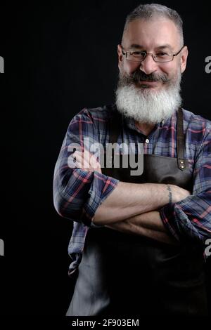 Ritratto di bel grembiule in pelle vestito uomo maturo isolato su sfondo nero, operaio caucasico con barba sorridente Foto Stock