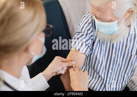 Visione offuscata di medico professionista sicuro in maschera facendo l'iniezione di vaccino o insulina al paziente anziano in maschera, visitandolo a casa. Sanità, vaccinazione, pandemia di covid-19. Foto Stock