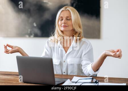 Rilassata tranquilla bella bionda senior fiduciosa, donna d'affari, in elegante camicia bianca, si siede sul posto di lavoro, ha preso una pausa dal lavoro, la meditazione, calma con gli occhi chiusi, calma e armonia Foto Stock