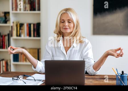 Pausa durante il lavoro. Bionda matura sicura, donna di affari o avvocato, indossando una camicia bianca, seduto ad una scrivania, ha preso una pausa dal lavoro del calcolatore, medita con gli occhi chiusi Foto Stock