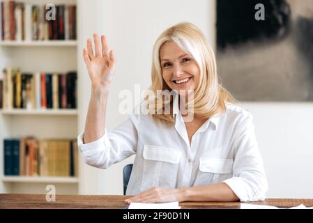 Intelligente bella donna bionda senior amichevole ceo o mentore di affari, chattando sulla comunicazione video con colleghi o clienti, guardando la fotocamera, accogliente con gesto della mano e sorridente Foto Stock