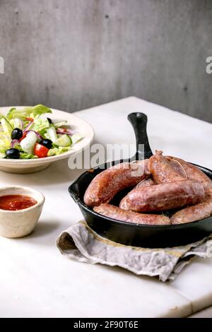 Salsiccia di salumi italiani alla griglia in padella di ghisa servita con salsa di pomodoro e piatto di insalata di verdure fresche su tavolo di marmo bianco. Cena bilanciata Foto Stock