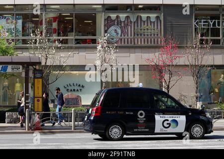 Una strada a Ginza con negozi, pedoni, alberi in fiore di ciliegio e un taxi Toyota JPN con il logo Tokyo Olympic 2020. (3/2021) Foto Stock