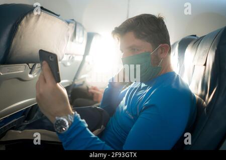 viaggi e turismo in tempi di covid19 - stile di vita ritratto Di giovane attraente uomo caucasico in maschera di faccia usando il mobile telefono in aereo durante il fligh Foto Stock