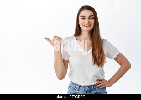 Sorridente allegra donna punta pollice a sinistra, mostra pubblicità copy space, negozio promo affare, guardare determinato, raccomandare prodotto, si trova contro bianco Foto Stock