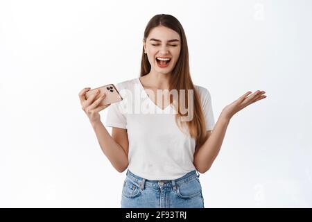 Felice giovane donna che gioisce, vincendo su smartphone, sorridendo e gridando sì con gioia, celebrando la vittoria online, tenendo il telefono orizzontale, vincere Foto Stock