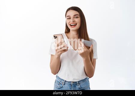 Donna allegra con smartphone e carta di credito. Felice ragazza che paga online senza contatto con il telefono, ridendo mentre lo shopping in app mobile, in piedi Foto Stock