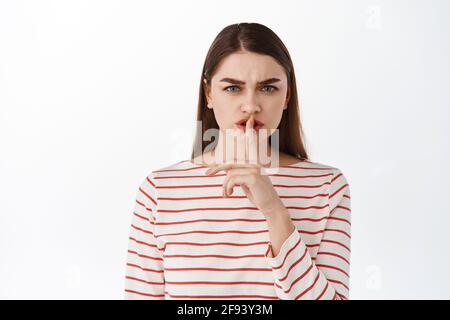 Fai silenzio. La ragazza spaventosa dice di chiudere, shushing, hushing con faccia accigliata arrabbiata, gesto tabù, non parlare, in piedi dispiaciuto contro bianco Foto Stock