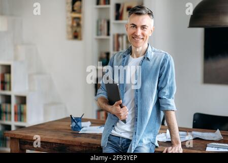 Ritratto maturo sicuro intelligente grigio-capelli caucasici uomo in abiti casual elegante, uomo d'affari o manager, si trova vicino al tavolo, tiene un portatile in mano, guardando la fotocamera, sorridente amichevole Foto Stock