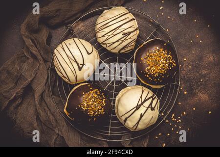 Pasticcini berlinesi tedeschi tradizionali smaltati con cioccolato bianco e scuro e fragola alle nocciole su sfondo marrone, vista dall'alto Foto Stock