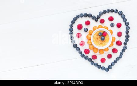 Simbolo del cuore fatto di bacche diverse con una creme brulee al centro su legno bianco, vista dall'alto con spazio per la copia Foto Stock