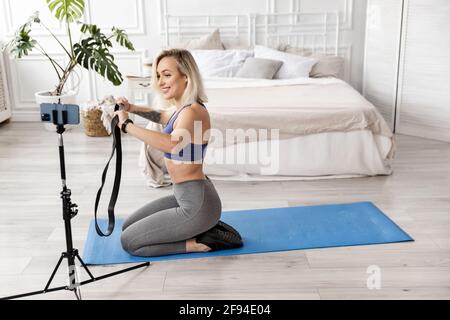 Donna sportiva felice sta usando la fascia di sport, facendo l'esercitazione per le armi, facendo l'addestramento in linea. Attraente allenatore femminile con smartphone Foto Stock