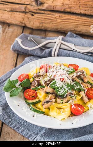 Cucina italiana: Tortellini con salsa di funghi vegetariani, zucchine e pomodori ciliegini freschi su un rustico tavolo in legno, verticale Foto Stock