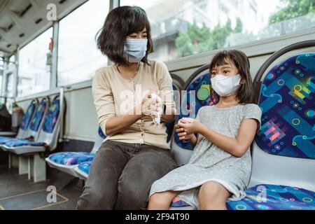 una madre indossa una maschera e sua figlia usa un tessuto per pulire le mani mentre si siede Foto Stock