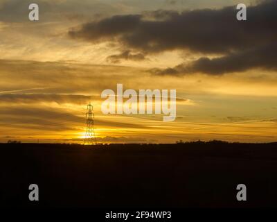 Un pilone elettrico, retroilluminato e silhouette dal sole che tramonta con un intricato paesaggio torbido stratificato in un enorme cielo dorato. Foto Stock