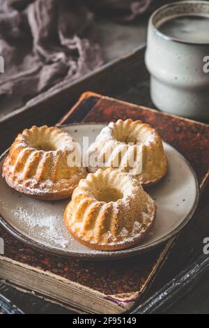 Tre dolci fatti in casa su un piatto, una tazza di caffè e un vecchio libro su un vassoio di legno su sfondo grigio, verticale Foto Stock