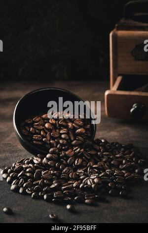 Chicchi di caffè che si inclinano verso il basso da una ciotola nera in primo piano, con il vecchio mulino vintage sullo sfondo. Foto Stock