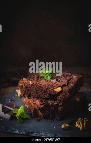 Pezzi impilati di brownie di noce su un piatto di ardesia e sfondo marrone scuro, decorati con noci, cioccolato e foglie di menta, verticali con copia Foto Stock