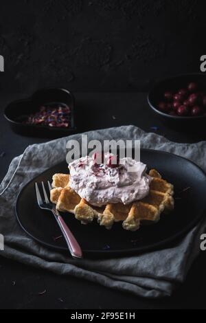 Waffle belgi con crema di mirtilli rossi, adagiati su un piatto nero e tovaglioli grigi, decorati con forchetta di rose e palline di cioccolato rosso Foto Stock