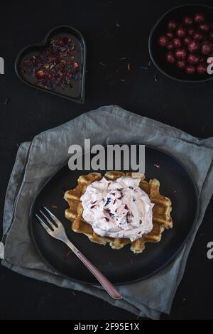 Waffle belgi con crema di mirtilli rossi, adagiati su un piatto nero e tovaglioli grigi, decorati con forchetta di rose e petali colorati e agili Foto Stock