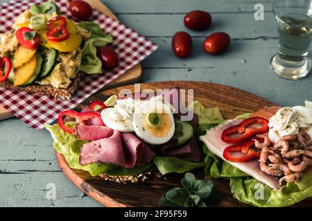 Varietà di panini danesi aperti con uova, frutti di mare e manzo arrosto, adagiati su tavole di legno su un tavolo di legno grigio chiaro. Foto Stock