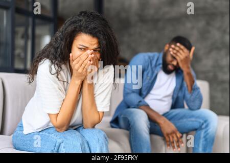 Concetto di problema di relazione di coppia biraciale giovane, donna africana che si sente triste e piangendo dopo avere un argomento a casa, donna triste o depressa seduta sul divano con la sua coppia, uomo in defocus si siede vicino Foto Stock