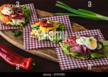 Varietà di panini danesi aperti con uova, frutti di mare e manzo arrosto, adagiati su tovaglioli controllati su una tavola di legno. Foto Stock