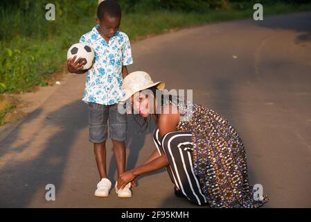 felice giovane donna che aiuta suo figlio a indossare sneakers Sul Road.sitting in piedi mentre tiene una palla di calcio Foto Stock