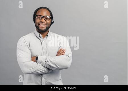 Amichevole e utile uomo afroamericano indossare cuffie e camicia casual intelligente guarda la fotocamera, sicuro ragazzo nero si alza con le braccia incrociate, addetto al servizio di assistenza in contatto, isolato su grigio Foto Stock